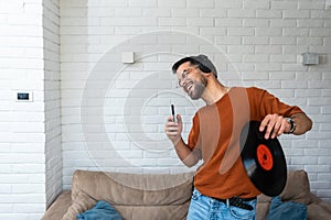 Young happy man singing and dancing at home listening music on wireless headset singing on his phone holding LP record in his hand