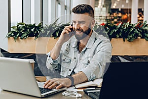 Young happy man sitting in office at table, working on computer, talking on phone.Freelancer has telephone conversations
