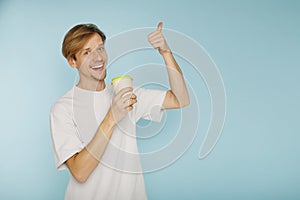 Young happy man standing over blue background smiling with happy face showing hand ok sign and drinking tea or coffee. Copy space