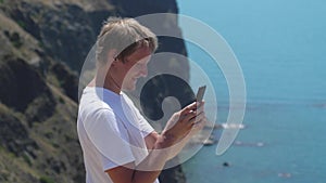 Young happy man taking photo with cellphone by the sea on the rock in slow motion. 3840x2160