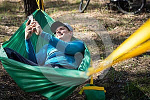 Young happy man using mobile phone in hat relaxing outside in hammock in forest.