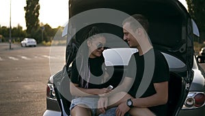 Young happy man and woman are sitting together in the open car trunk. Smiling, joyful couple caress each other, laughing