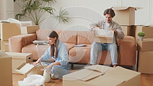 Young happy man and woman unpacking crockery from moving boxes, sitting at new own apartment, slow motion