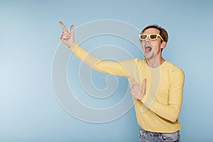 Young happy man in yellow sunglasses standing over blue background smiling with happy face looking and pointing to the side with