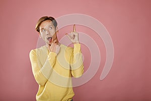 Young happy man in yellow sweater standing over pink background with shocked face looking and pointing to the side with thumb up.