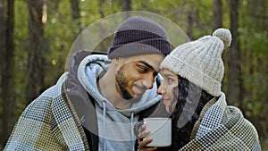 Young happy married couple on date in nature guy and girl lovers in autumn forest embracing in warm blanket drinking tea