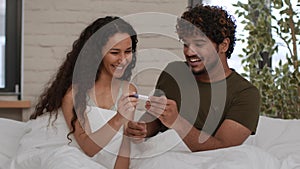 Young happy middle eastern man and woman looking at positive pregnancy test result and laughing, enjoying moment in bed