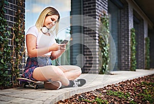Young happy millennial woman sitting in front of a building with headphones on her neck - Teenager skateboarder girl using
