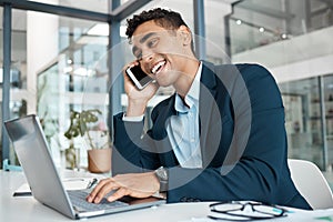 Young happy mixed race businessman talking and laughing on a call using a phone and typing on a laptop alone at work