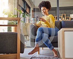 Young happy mixed race woman drinking a cup of coffee and typing a message on a phone at home. One content hispanic