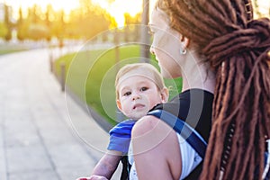 Young happy mom with baby son in ergo backpack travel together. Sunny summer day in the Park. Concept of modern parents