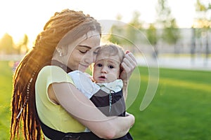 Young happy mom with baby son in ergo backpack walking in the Park. Sunny summer day. Concept of modern parents