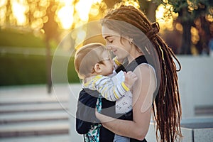 Young happy mother with baby son in ergo backpack walking in Sunny summer day. Concept of the joy of motherhood