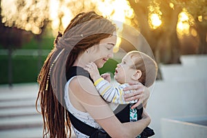 Young happy mother with baby son in ergo backpack walking in Sunny summer day. Concept of the joy of motherhood