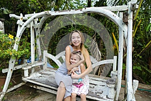 Young happy mother with daughter riding on swing in exotic garden, palms in background.