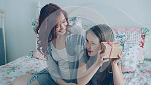 Young happy mother giving gift box to her little cute daughter celebrating birthday sitting on bed in cozy bedroom at