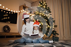 Young happy mother and little cute daughter in santa hat reading magical fairy tale book