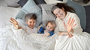 Young happy mother lying in bed with her two sons and waving in camera. Concept of family happiness, relaxing at home, having fun