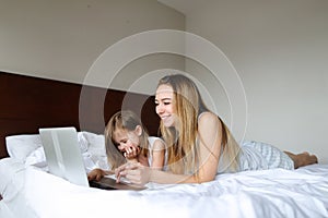 Young happy mother lying in bed with little daughter and using laptop.