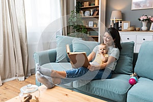 Young happy mother reading a fairytale story book to her baby. Mommy and kid sitting on sofa at home enjoying in imagination.