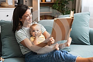 Young happy mother reading a fairytale story book to her baby. Mommy and kid sitting on sofa at home enjoying in imagination.
