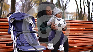 Young happy mother sitting with toddler son on bench in autumn park talking playing with soccer ball. Portrait of