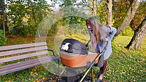 Young happy mother stands with a stroller in the park, smiles at the baby and takes the baby out of the stroller. Full