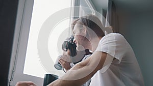 Young happy multiracial couple drinking coffee or tea and looking through the window. Slow motion