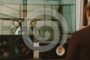 young happy musicians recording song behind glass at studio and showing