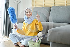 Young happy muslim woman wearing yellow gloves  and holding a basket of cleaning supplies in living room