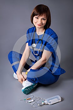Young happy nurse sitting on the floor posing on gray background. Medical dropper and a stethoscope of a medic