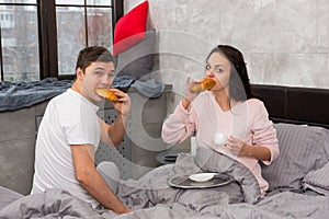 Young happy pair just woke up, eating croissants and having breakfast in bed