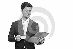 Young happy Persian businessman reading clipboard while holding coffee cup