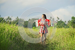 Young happy and playful Asian Chinese woman in beautiful dress having fun enjoying holidays excursion on grass tropical field smil