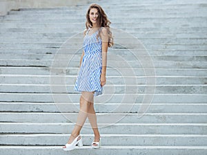 Young happy playful woman in light striped white blue dress posing at concrete stairway outdoor