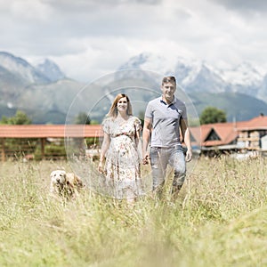 Young happy pregnant couple holding hands walking it`s Golden retriever dog outdoors in meadow.