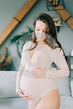 A young happy pregnant woman in a beige bodysuit is standing in a cozy spacious apartment, smiling and holding her stomach