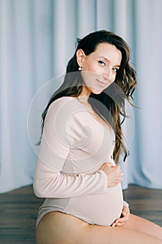 A young happy pregnant woman in a beige bodysuit stands next to a blue fabric background, smiling and holding her stomach