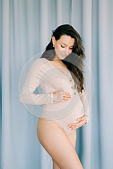 A young happy pregnant woman in a beige bodysuit stands next to a blue fabric background, smiling and holding her stomach