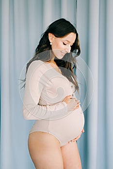 A young happy pregnant woman in a beige bodysuit stands next to a blue fabric background, smiling and holding her stomach