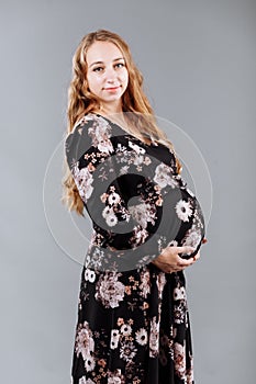 A young happy pregnant woman in an elegant floral dress standing with hands on her belly in front of a grey background in a studio