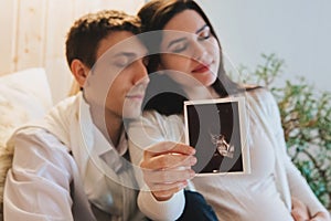 Young happy pregnant woman holding pregnancy ultrasound, showing sonogram picture to pleasantly surprised husband crying from