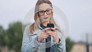 Young happy pretty smiling hipster girl in glasses using modern smartphone outdoors, beautiful woman texting sms message