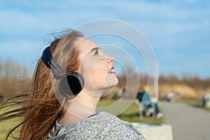 Young, happy redhead girl in the spring in the park near the river listens to music through wireless bluetooth headphones