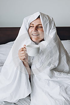 Young happy redhead woman with cup of coffee sitting in bed under blanket at home bedroom. comfort or home weeekend