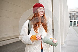 Young happy redhead woman in red hat, drinking from thermos, warming up with hot drink in her flask while walking around