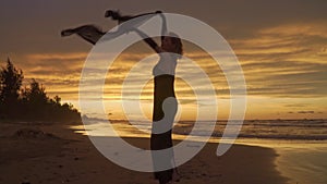 Young happy romantic girl is standing or dancing at sunset beach with raised arms and using a scarf.