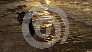 Young happy romantic girl is standing or dancing at sunset beach with raised arms and using a scarf. Full length