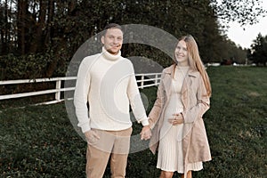 Young happy romantic pregnant couple is walking holding hands outdoors at autumn warm day. Pregnant woman in knit dress