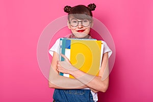 Young happy schoolgirl embraces colorful binders isolated on pink background. Smiling girl looks happy, passes final exames, ready